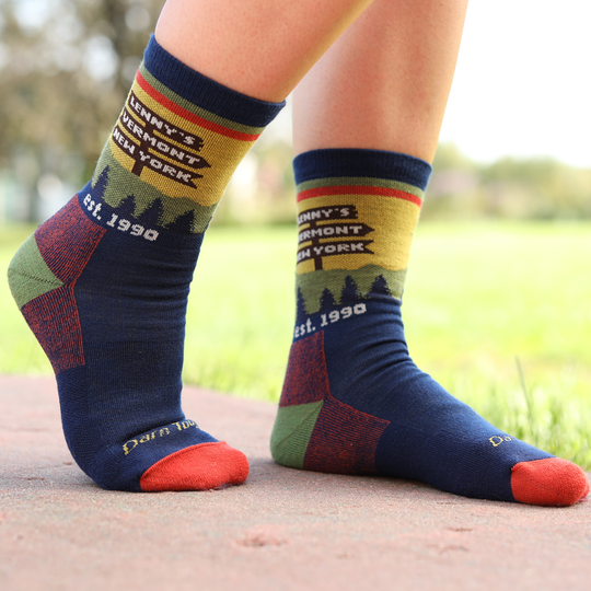 A close-up of a person's feet wearing colorful crew socks on an outdoor path with grass in the background. The socks are navy blue with red, green, and yellow accents, featuring a graphic of a mountain skyline and the text 'Lenny's New York', 'Est.