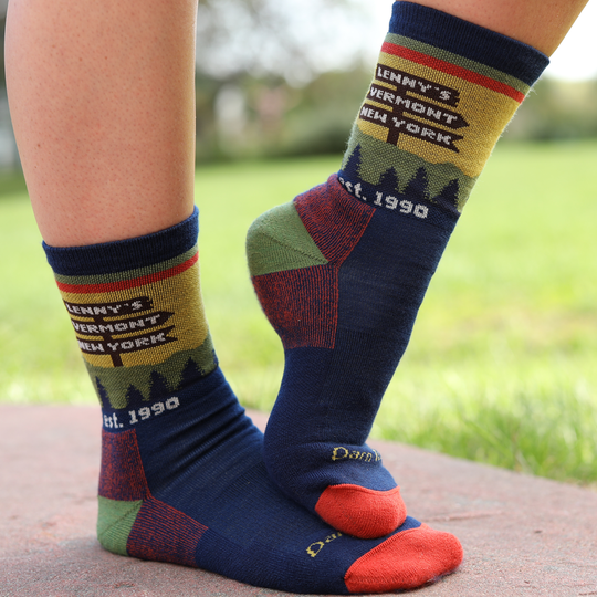 A person wearing a pair of crew socks outdoors on a stone path with grass in the background. The socks are navy blue with olive green, red, and tan sections, featuring printed text that reads 'LENNY'S VERMONT NEW YORK est.