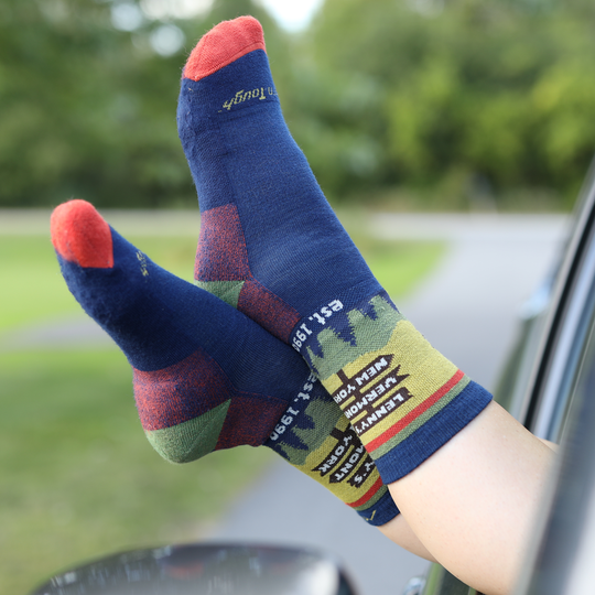 A pair of crew socks worn on feet sticking out of a car window, with a blurred outdoor background. The socks are navy blue with red toes, green and red geometric panels, and a yellow-green lower section featuring a forest pattern and directional sign graphic.