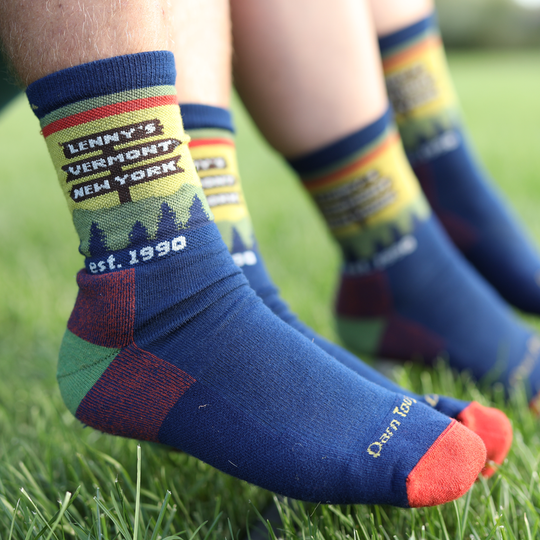 Close-up of several legs sitting on grass wearing matching blue crew socks with colorful printed graphics. The front sock has a sign reading "Lenny's Vermont New York" and "est.