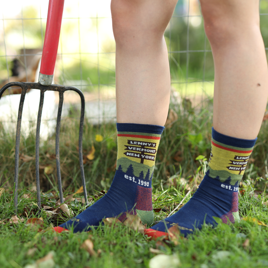 A person standing outdoors on grass wearing patterned crew socks with a red-handled garden fork visible behind them. The socks are navy blue with a multicolor graphic design and text reading 'Lennys Version New York est.