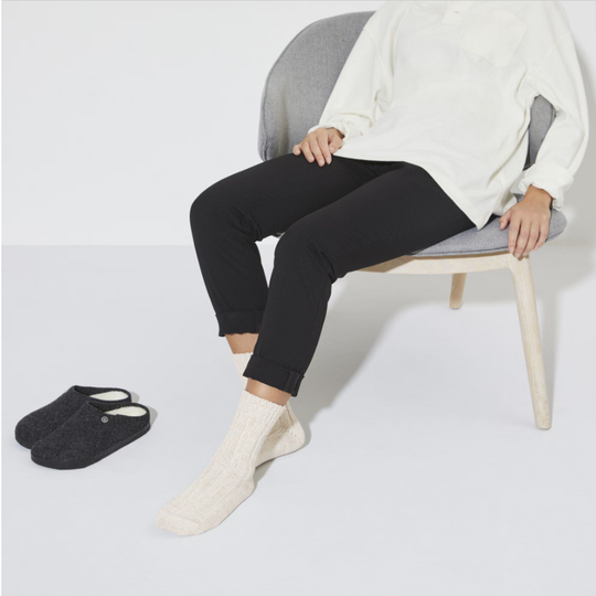 Person sitting on a chair wearing black pants, white socks, and a white sweater with a pair of black slippers on the floor.