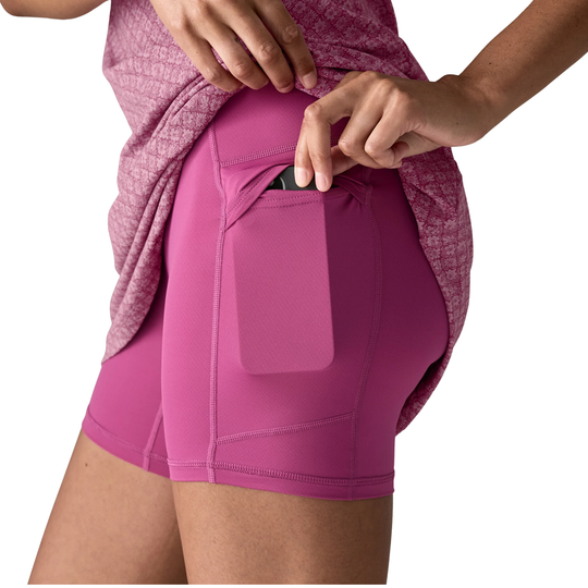 A close-up of a person wearing bright pink athletic shorts with a side pocket, pulling a phone out of the pocket while wearing a matching heathered pink top against a white background.