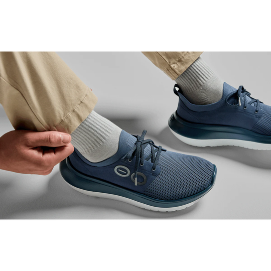 A close-up of a person wearing a navy blue mesh athletic sneaker with a white sole, paired with light gray socks and khaki pants. The person's hand is adjusting the pant leg, and the shoe features a visible circular logo on the side.