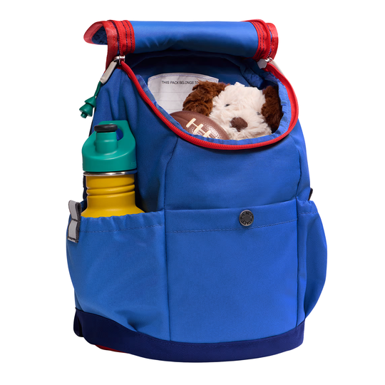 A blue soft-sided backpack or pet carrier shown on a white background, with red trim, a front pocket, side pockets, a green-topped bottle in a side pocket, and a stuffed dog toy visible in the top opening.