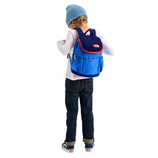 A child wearing a light blue knit beanie, white long-sleeve shirt, dark blue jeans, and blue sneakers, shown from the back while wearing a blue and navy backpack with red trim. The child is photographed against a plain white background.