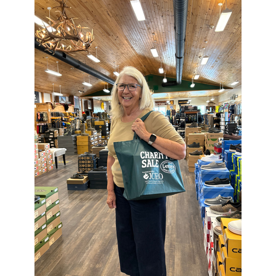 A smiling blonde woman wearing glasses stands inside a retail store holding a teal tote bag printed with 'CHARITY SALE' and the JCPen brand logo. The store has a wood ceiling, chandeliers, and shelves of products.