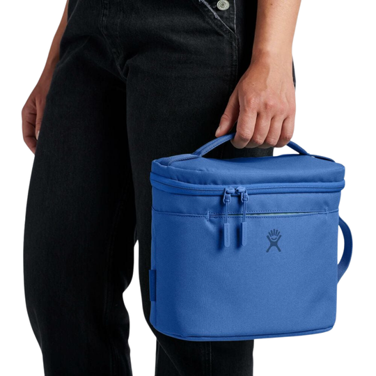 A person wearing black jeans holds a bright blue insulated lunch bag with a top handle and side loop, shown against a plain white background. The bag has a dark logo on the front and a zipper closure.