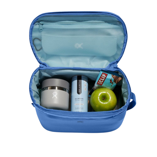 An open blue lunch bag shown from above on a white background, containing a gray insulated tumbler, a light blue bottle, a green apple, and a snack bar.