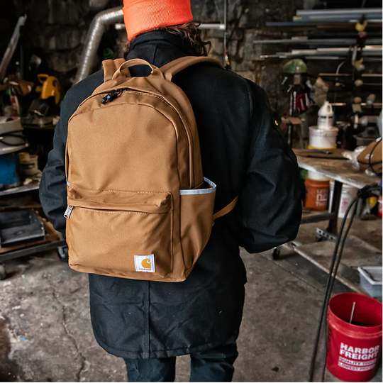 A person viewed from behind wearing a tan Carhartt backpack in a workshop environment. The person is also wearing a black work jacket, dark pants, and an orange beanie.
