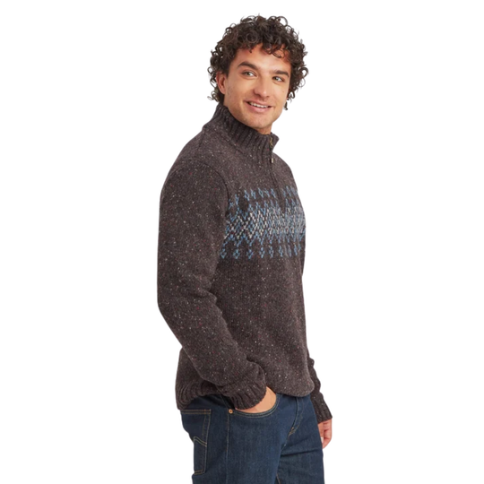 A man wearing a dark brown knit turtleneck sweater with a blue and gray geometric pattern across the chest, paired with blue jeans, photographed against a solid white background.