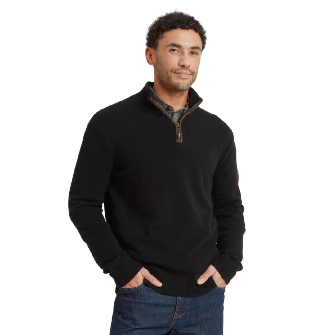 A man wearing a black quarter-zip knit sweater over a plaid collared shirt and blue jeans, standing against a plain white background.