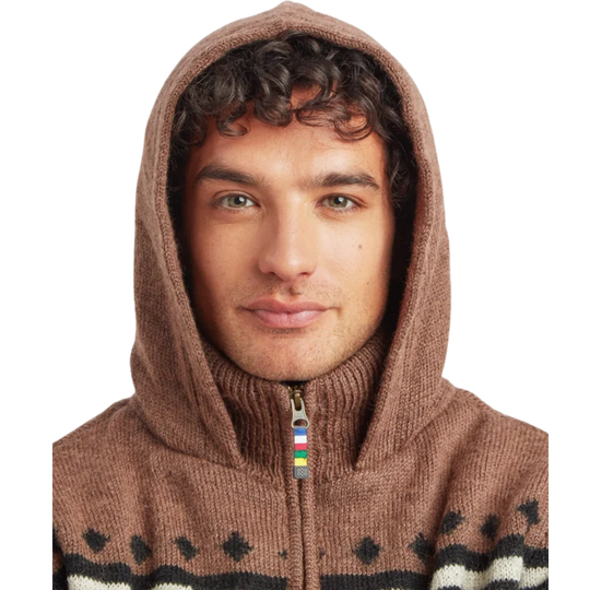 A close-up of a man wearing a brown knit zip-up hoodie with a high collar, black and white geometric patterns along the hem, and a small rainbow-colored zipper pull.