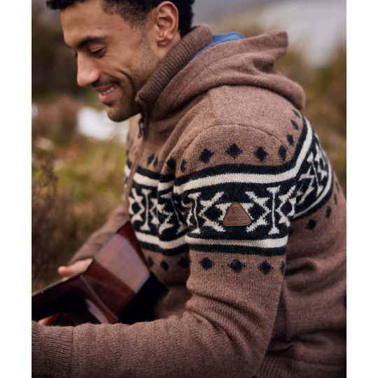 A man wearing a brown knit hooded sweater with a black and cream geometric pattern, holding an acoustic guitar in an outdoor setting.