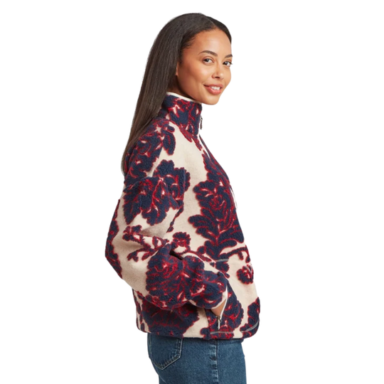A woman wearing a quarter-zip fleece pullover with a bold floral pattern in navy blue and red on a cream background, paired with blue jeans, shown from a side-back profile against a white studio background.