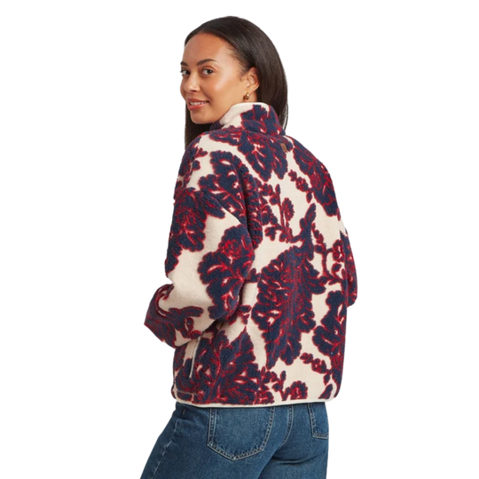A woman wearing a quarter-zip fleece pullover with a bold floral pattern in navy blue and red on a cream background, paired with blue jeans, shown from a side-back profile against a white studio background.