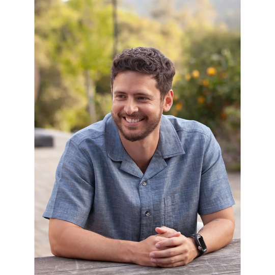 A smiling man with short curly brown hair and a beard, wearing a textured blue short-sleeve button-up shirt and a black smartwatch, leaning on a wooden table outdoors with a blurred garden background.