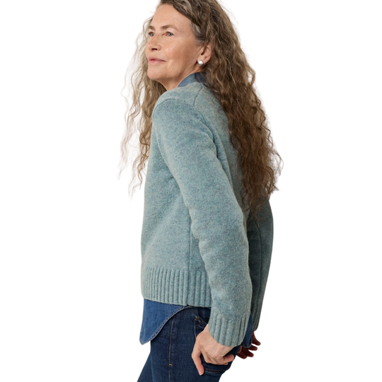 A woman with long curly brown hair is shown from a side-back angle wearing a heathered sage green knit cardigan over a blue shirt and dark jeans, against a plain white background.