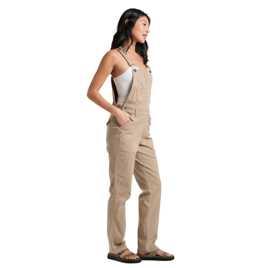 A woman modeling a pair of light tan canvas bib overalls with front pockets, worn over a white tank top and black sandals, shown from a side profile against a solid white background.