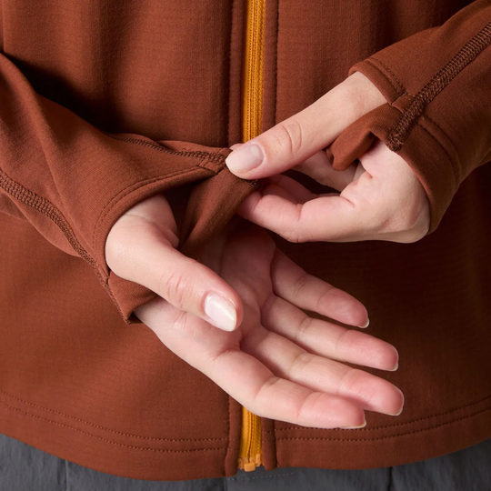Close-up of a person's hands adjusting the cuff of a rust-brown zip-up jacket, showing the yellow zipper and fabric texture.
