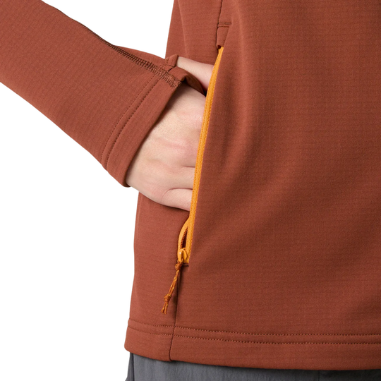 Close-up of a person wearing a rust-orange long-sleeve top, showing a side pocket with a yellow-orange zipper and drawstring being pulled open by a hand.