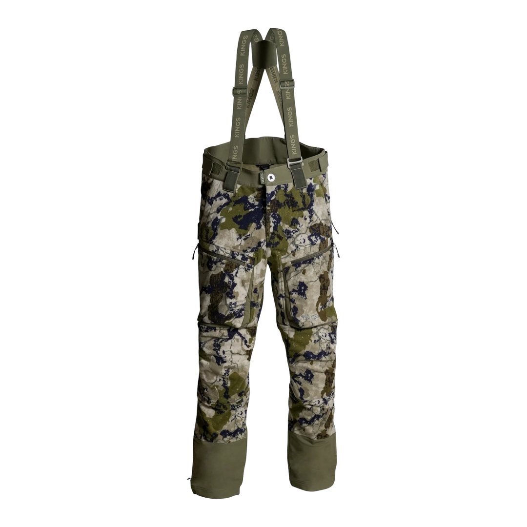 A pair of camouflage bib overalls with olive green suspenders, featuring a digital camo pattern in beige, green, and black, with solid olive green reinforced cuffs.