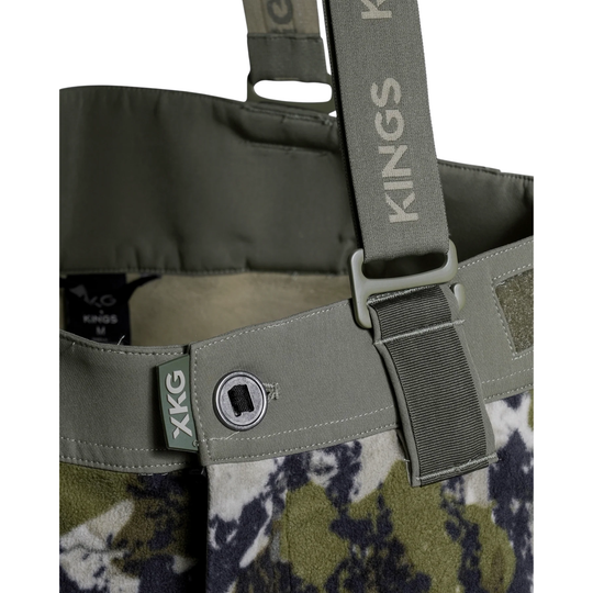 Close-up of a KXG Kings Waders chest panel in olive green nylon webbing with a metal snap closure, attached to a green and navy camouflage lower body panel.