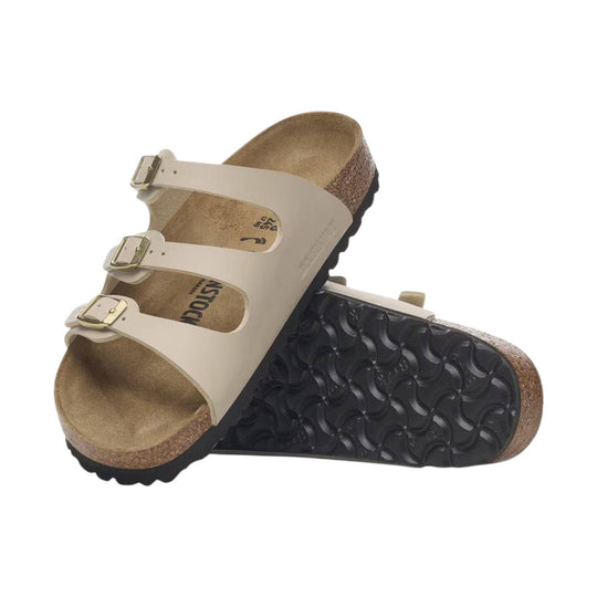 Beige triple-strap sandal with gold buckles, cork footbed, and black treaded outsole shown stacked on a white background