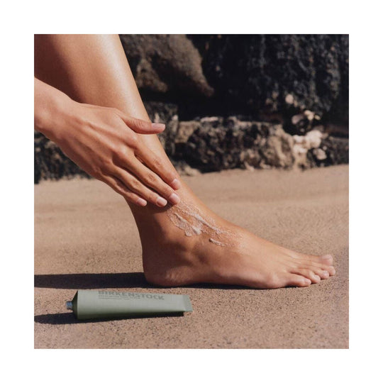 Foot cream tube beside a bare foot being moisturized on sand