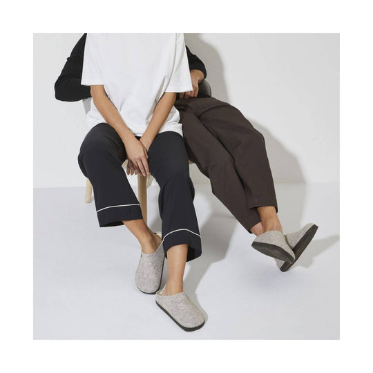 Two models seated wearing slip-on clog slippers, one light gray and one dark brown