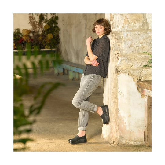 Black platform clogs with stitched sole worn by a model leaning against a stone wall