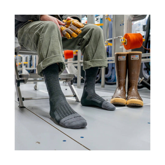 Brown rubber work boots with tan soles beside a seated person wearing gray work socks