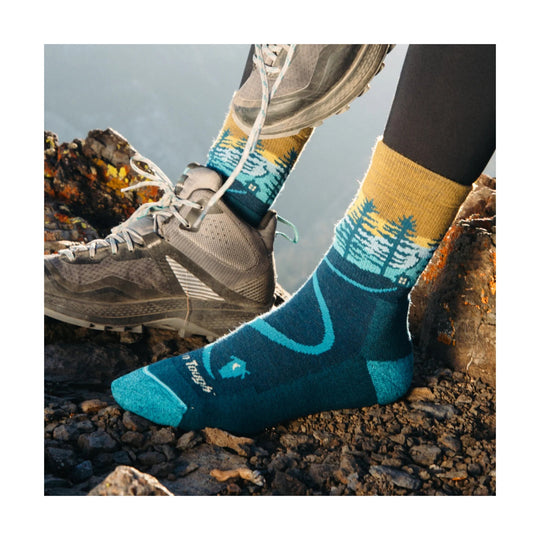 Blue and teal hiking sock with mountain pattern, shown with gray trail shoes on rocky ground