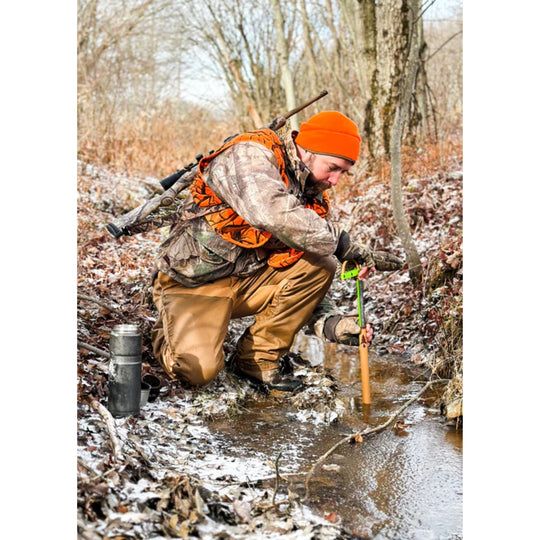 Hunter kneeling by a stream in camouflage gear with an orange vest and beanie, using a water filter