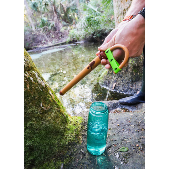 Hand using a tan and green water filter straw to fill a blue bottle from a stream