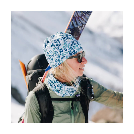 Floral blue-and-white beanie and neck gaiter worn by a skier with sunglasses and skis on her back