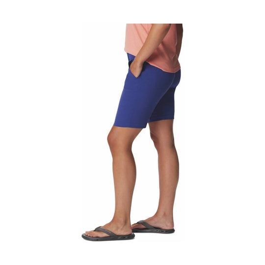 Woman wearing blue athletic shorts with hand pockets and gray flip-flops, shown in side view